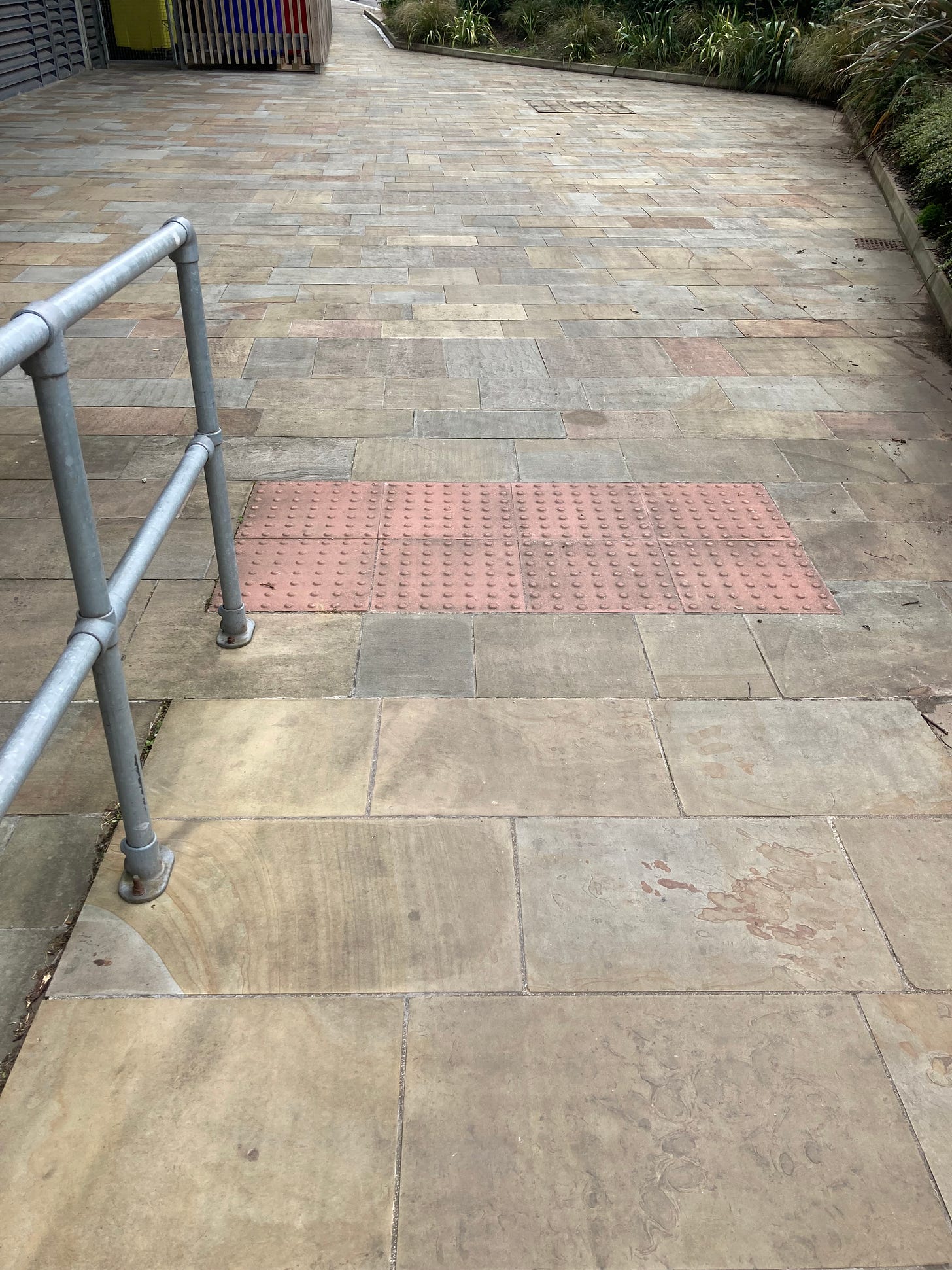 Ramp in completely pedestrianised area, where no cars can access. At the bottom of the ramp, there is a strip of red blister paving for no apparent reason. Ramp in completely pedestrianised area, where no cars can access. At the bottom of the ramp, there is a strip of red blister paving for no apparent reason.