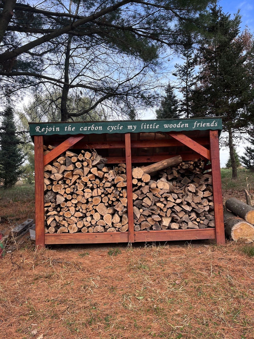 A wooden shed stacked with firewood under a sign reading "Rejoin the carbon cycle my little wooden friends."