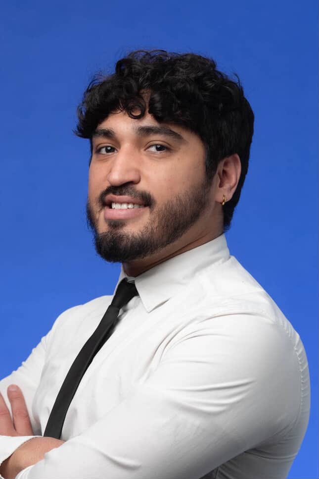 Cristino N. Chávez Jr. (left image): Cristino N. Chávez poses confidently against a solid blue background. He is wearing a white dress shirt with a black tie and has his arms crossed. He has short curly dark hair, a trimmed beard, and a small hoop earring.  Pius Semutumba (right image): Pius Semutumba takes a selfie outdoors near a university campus. He is wearing a gray knit hat, a black jacket, and a red backpack. The sky is partly cloudy, and campus buildings with glass and white walls are visible in the background.