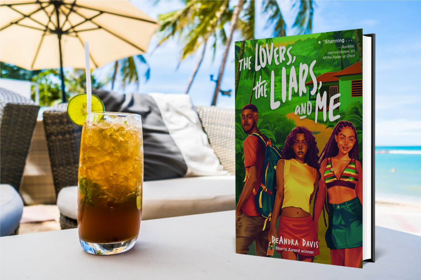 book "The Lovers, the Liars, and Me" next to a drink on a table by the beach