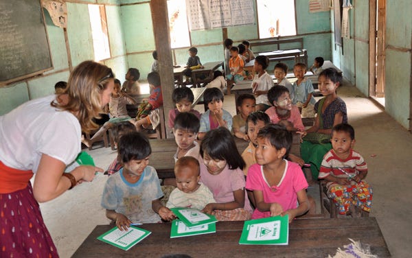 A group of kids sitting behind wooden desks with a white woman with her back to the camera, passing out green notebooks to them.