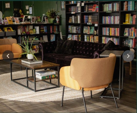 a neon, pastel sign with a rainbow and cursive writing, hung above a table with merchandise, says "The Future is Inclusive." A yellow chair and couch encircle a coffee table with a plant on top, creating an inviting gathering space..