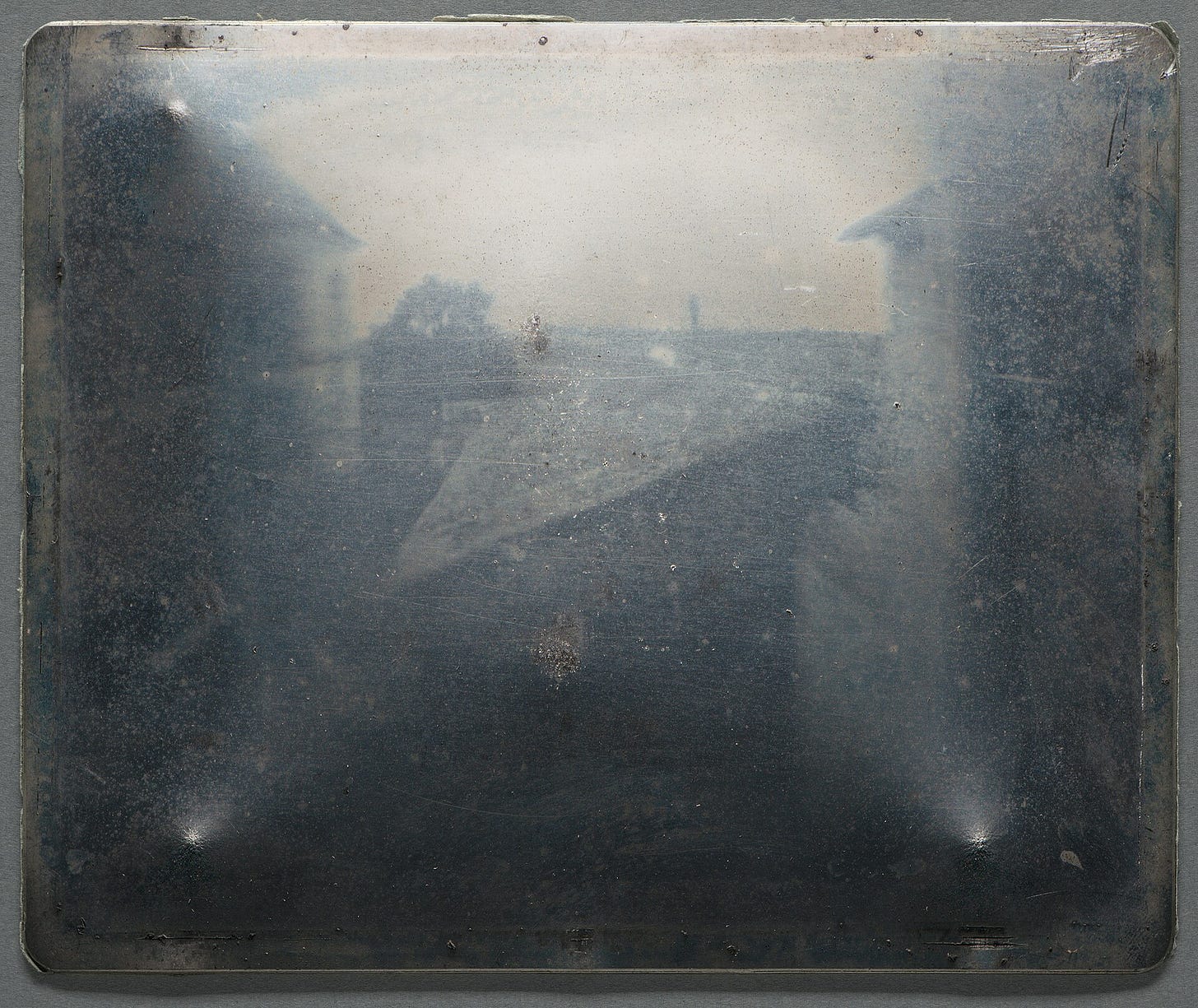 A faint image on a metal (pewter) backing. The metal is scuffed, but some rooftops, the top of a tree and the horizon are visible.