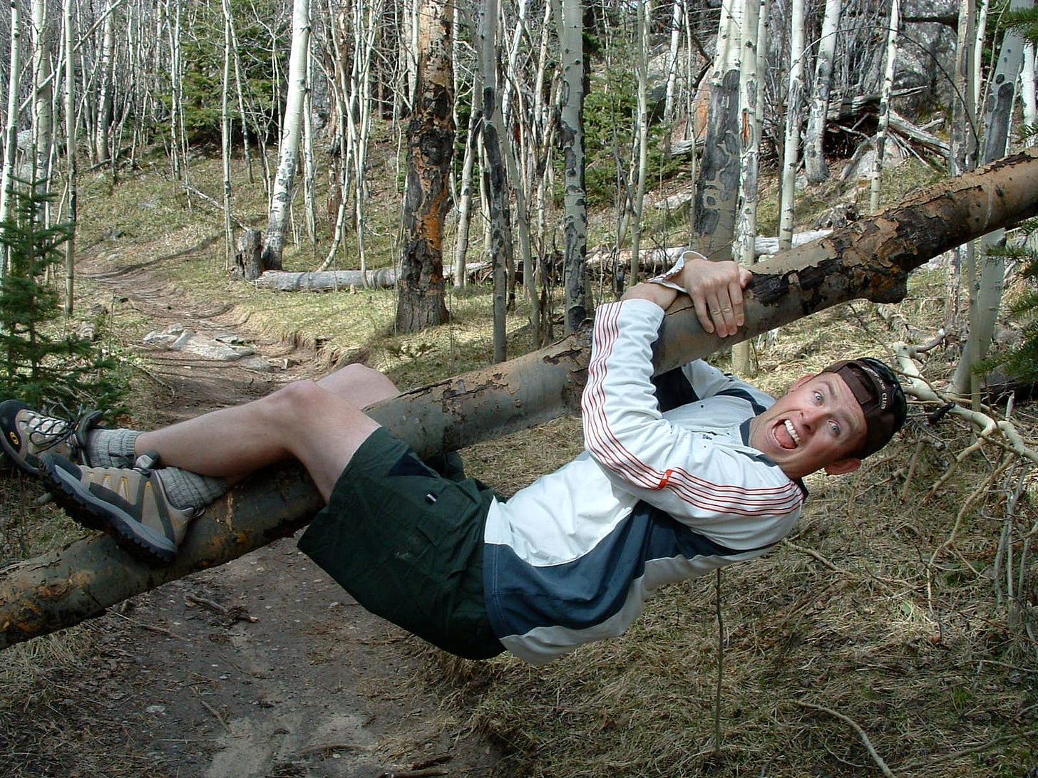 Kevin hanging sideways from a fallen tree in the woods, mid-laugh, legs wrapped around the trunk — a snapshot of pure, ridiculous joy in nature.