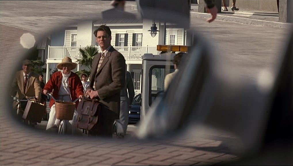 Truman stands in a bright, orderly town, seen through a distorted circular frame that suggests the perspective of a hidden camera.