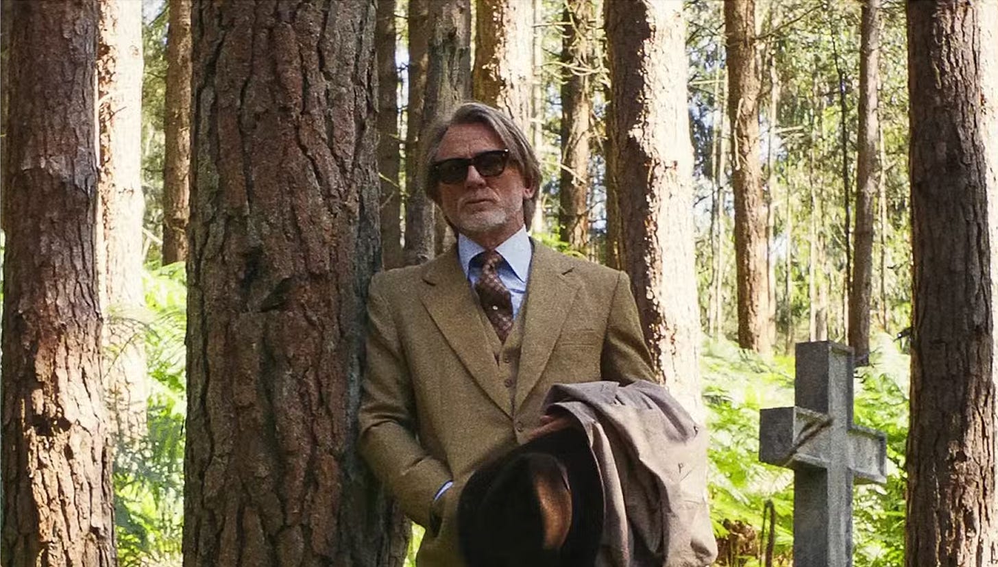 Daniel Craig with sunglasses holding a wide-brim hat leaning against a tree in a forest; a cross for a grave is off to his side and slightly behind him.