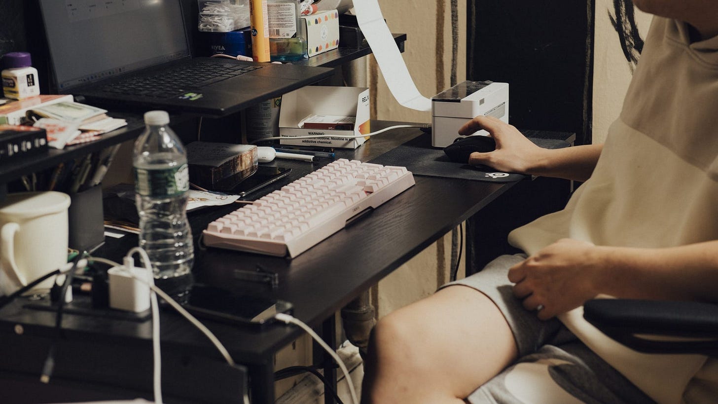 A person sitting at a desk with a pink keyboard and a computer mouse, using a laptop while surrounded by various items, a roll of paper used to print packaging labels is in focus.