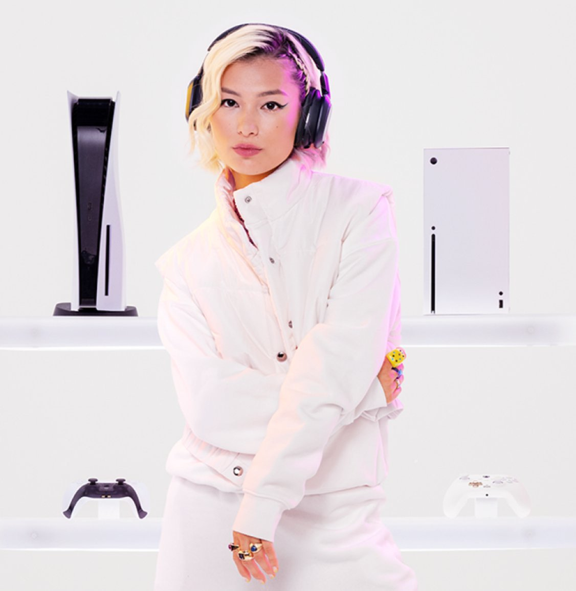 A white Xbox Series X and PS5 in the background of an Asian woman wearing headphones dressed in white A white Xbox Series X and PS5 in the background of an Asian woman wearing headphones dressed in white