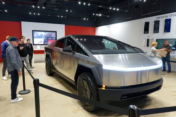 A Tesla Cybertruck on display at a showroom. The truck is behind a tape barrier. A group of people on the left are looking at the vehicle.