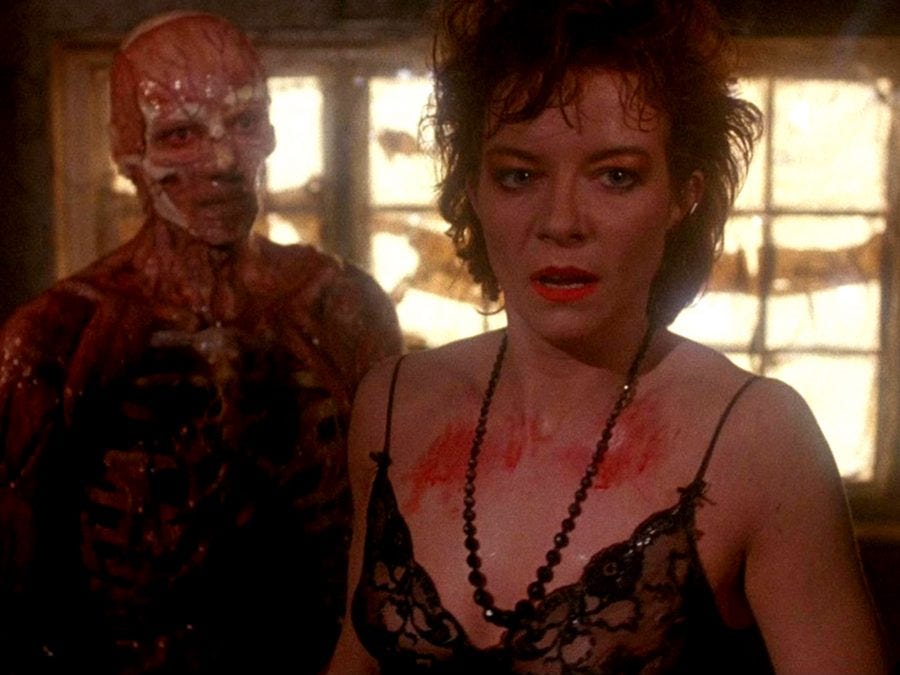 A medium shot of a flayed human body standing behind a white woman wearing a black negligee, with someone else's blood on her chest