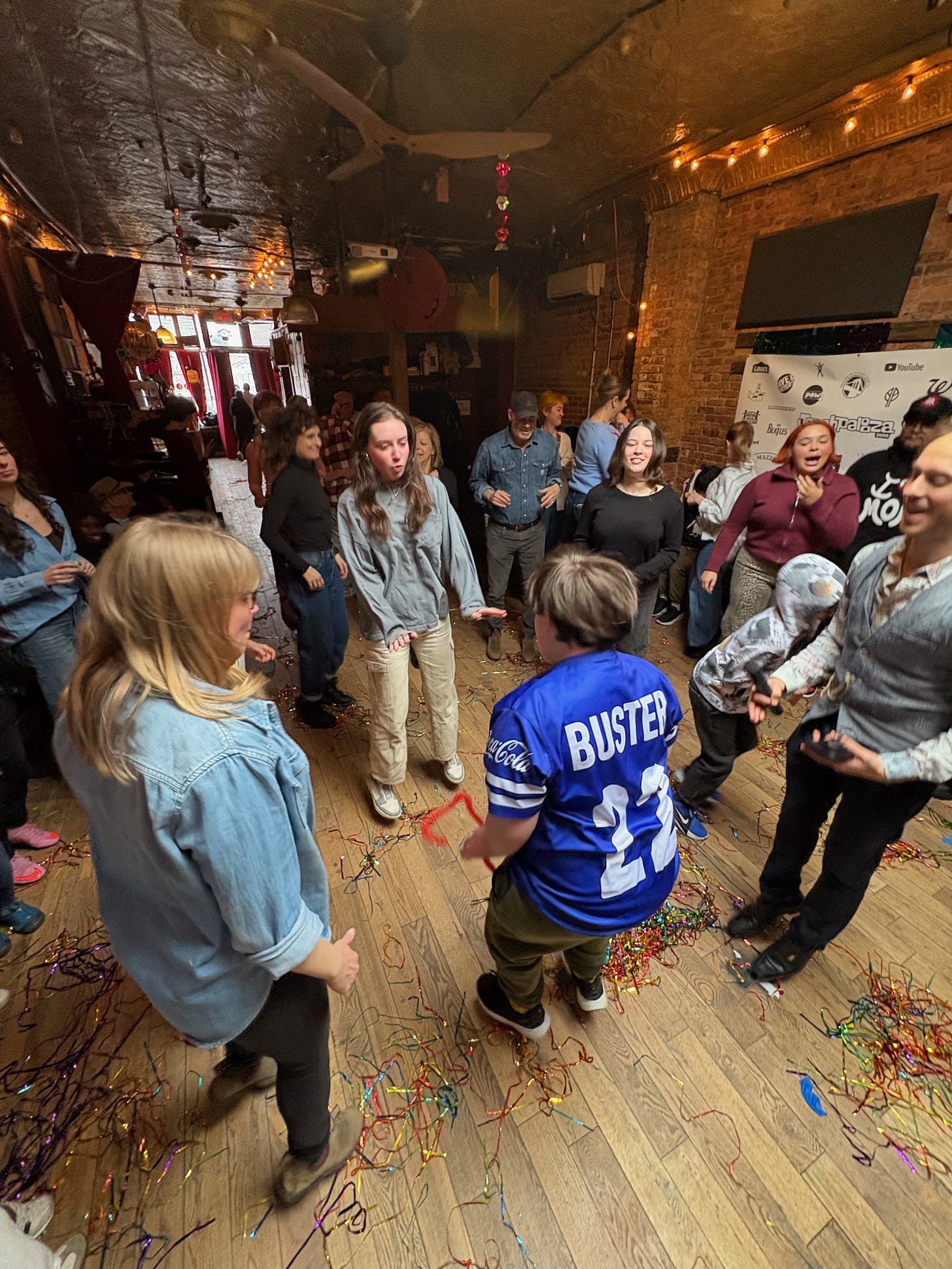 A group of people stand in a circle, dancing together in a room with brick walls and a wooden dance floor strewn with colorful tinsel  