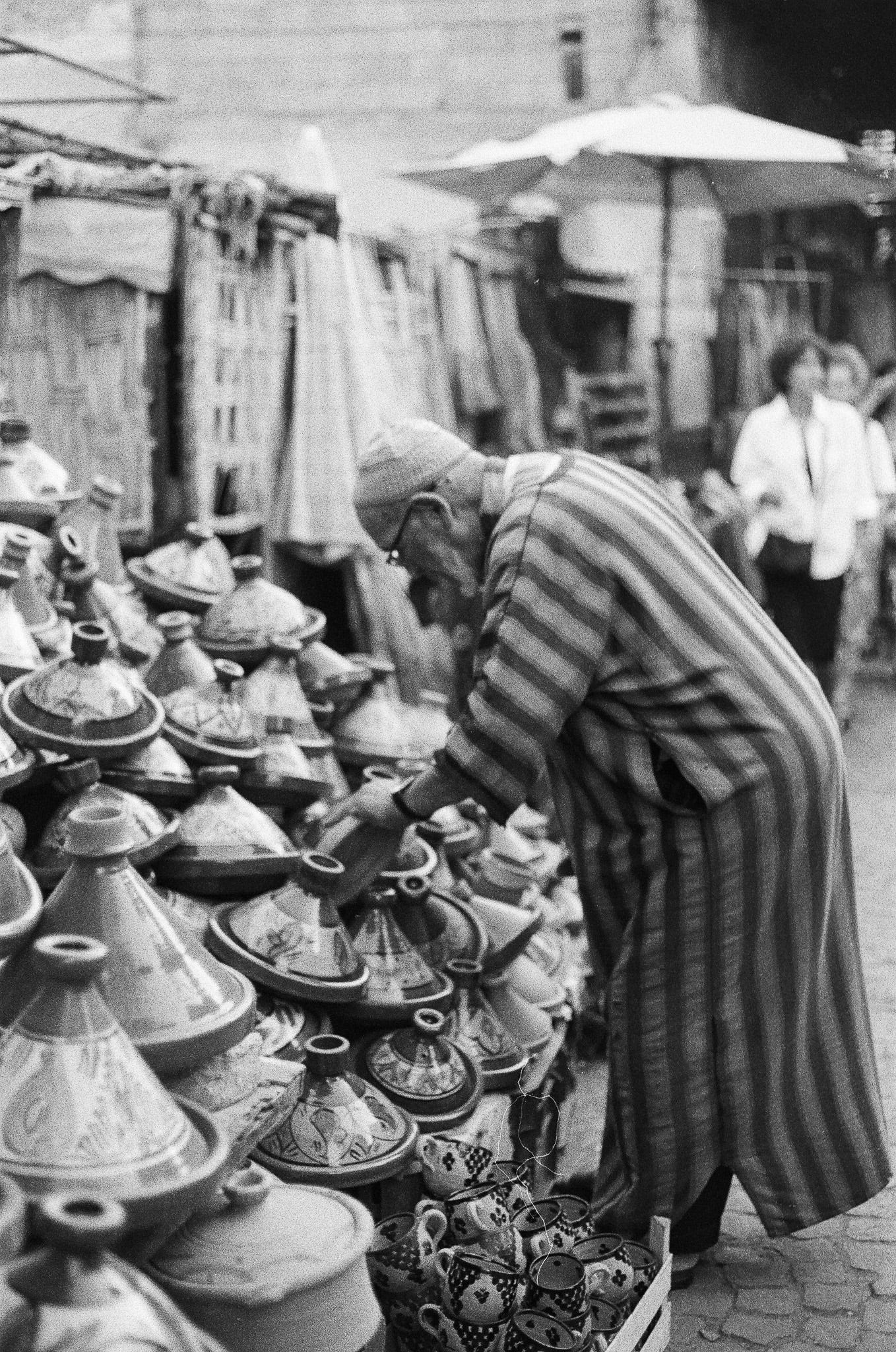 Black-and-white photograph of an older man in a striped djellaba sorting ceramic tagine pots at a market stall in Marrakesh.