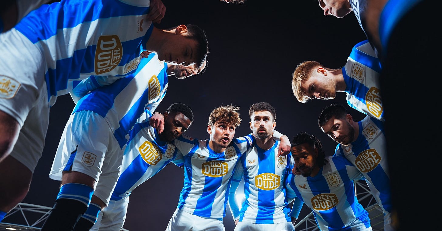 Huddersfield Town players huddle together on the pitch before a match, shot from below looking up, with the blue and white striped home kit and sponsor visible.​​​​​​​​​​​​​​​​