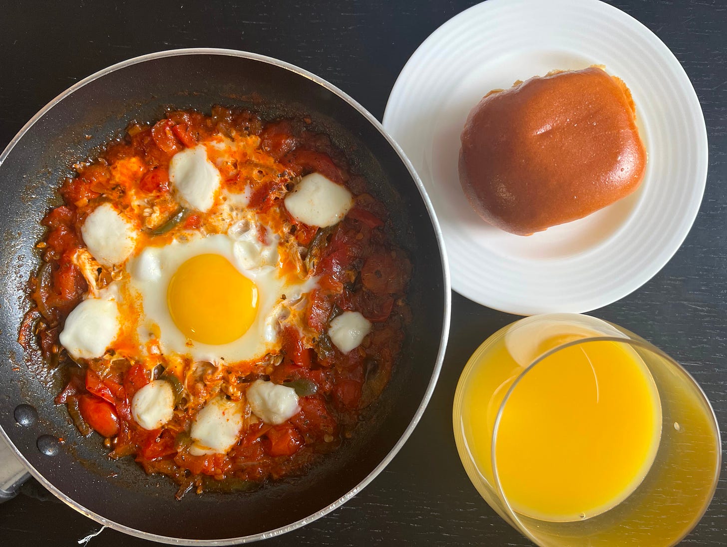 A skillet of shakshuka topped with fresh mozzarella chunks, baked to perfection with a golden yolk in the center, accompanied by a soft, glossy brioche roll and a glass of orange juice. A bold fusion of flavors, blending traditional Israeli shakshuka with modern influences. Another creation by Liat Portal for The Liat Show and Foodie Disorder.