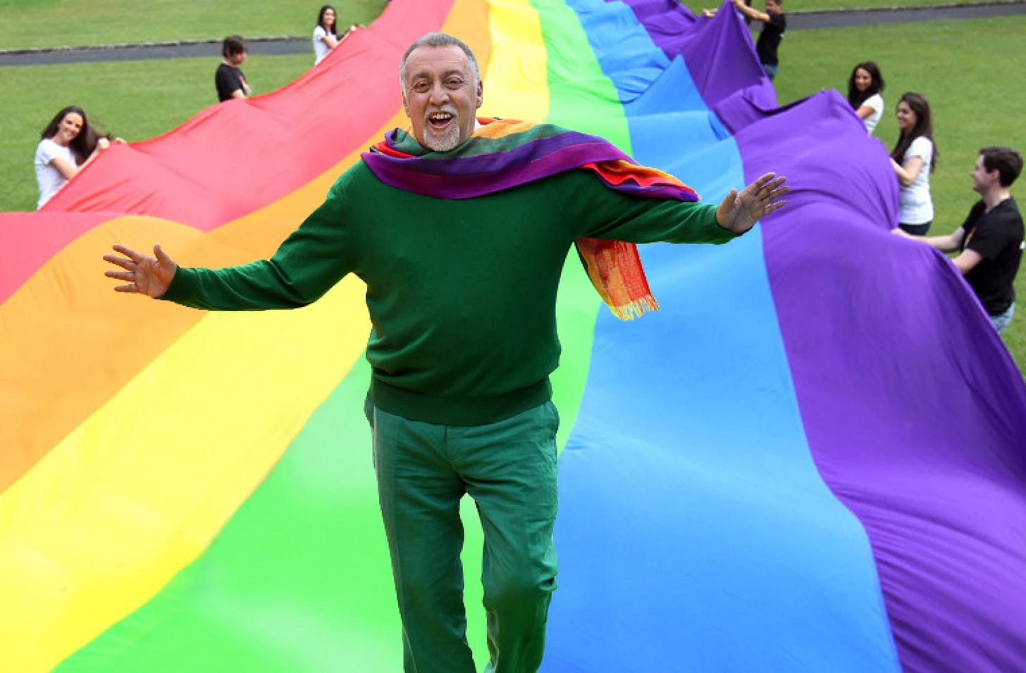 A man dressed in green stands in front of a giant rainbow flag. A man dressed in green stands in front of a giant rainbow flag.