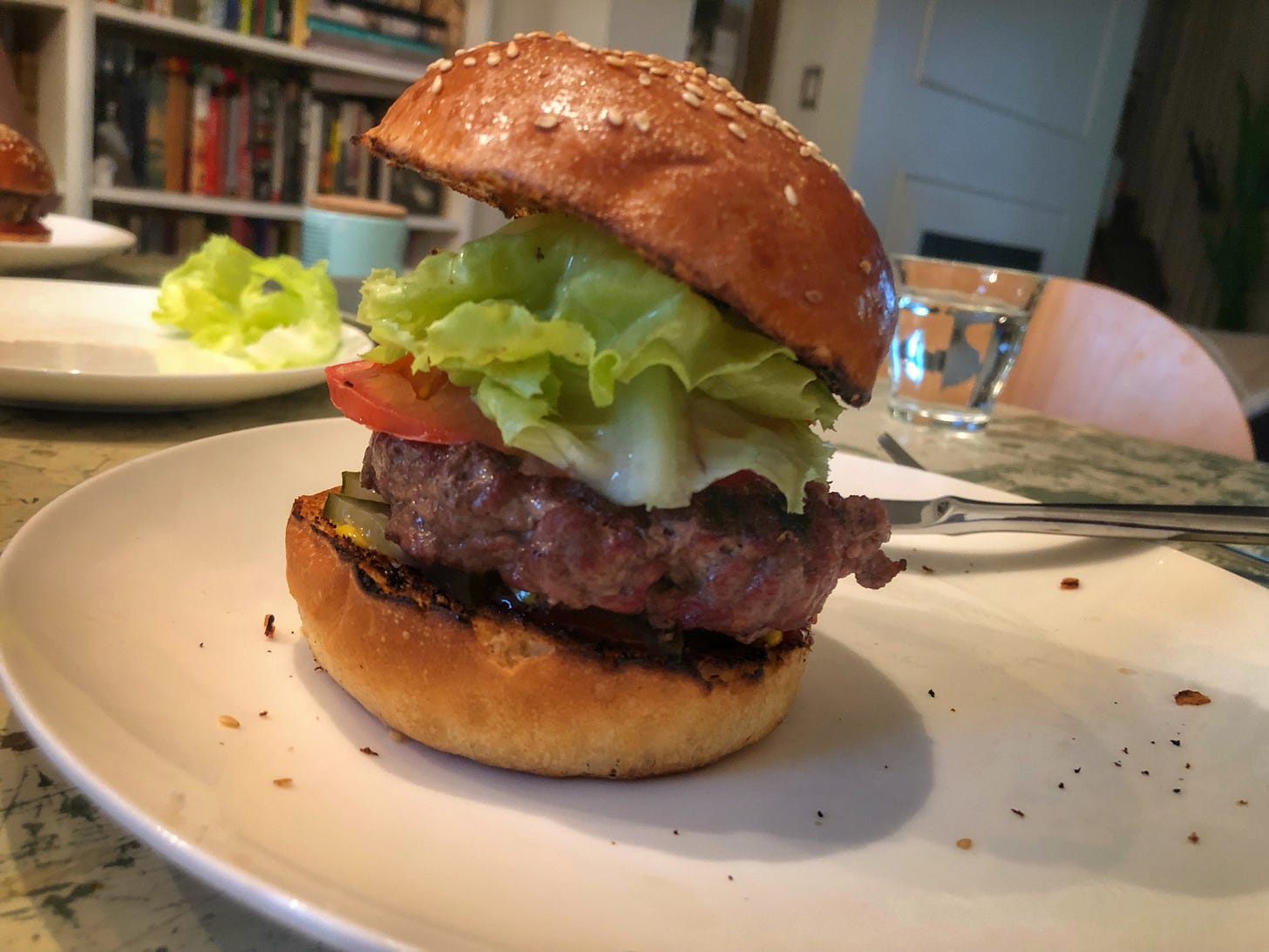 A dinner plate with a burger on a sesame seed bun, piled high with lettuce and tomato. It is far too tall to fit into someone's mouth.