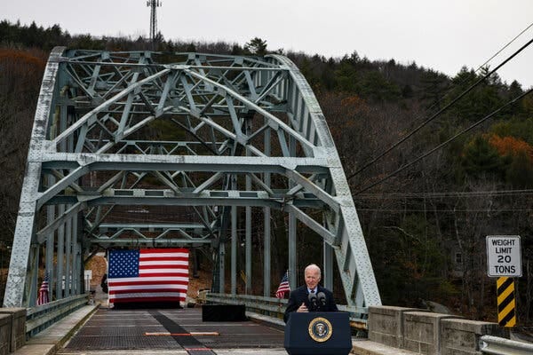Joe Biden speaks at a podium set up at a bridge, where an American flag is hanging.