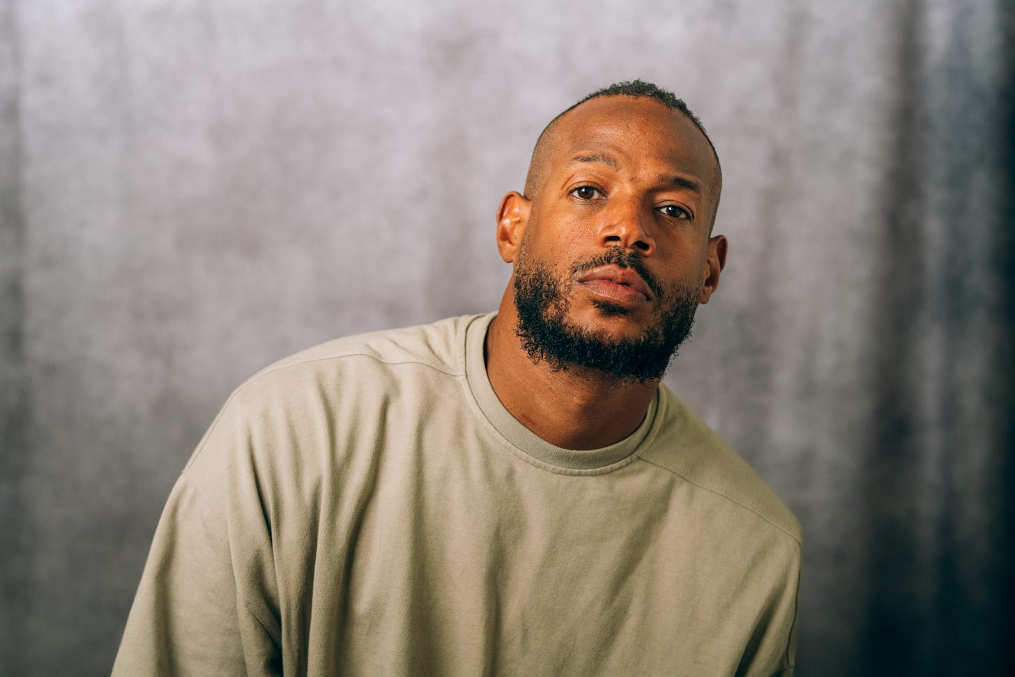Marlon Wayans leans slightly forward, wearing a beige crewneck shirt, gazing intently into the camera with a soft, contemplative expression against a neutral, softly blurred backdrop.
