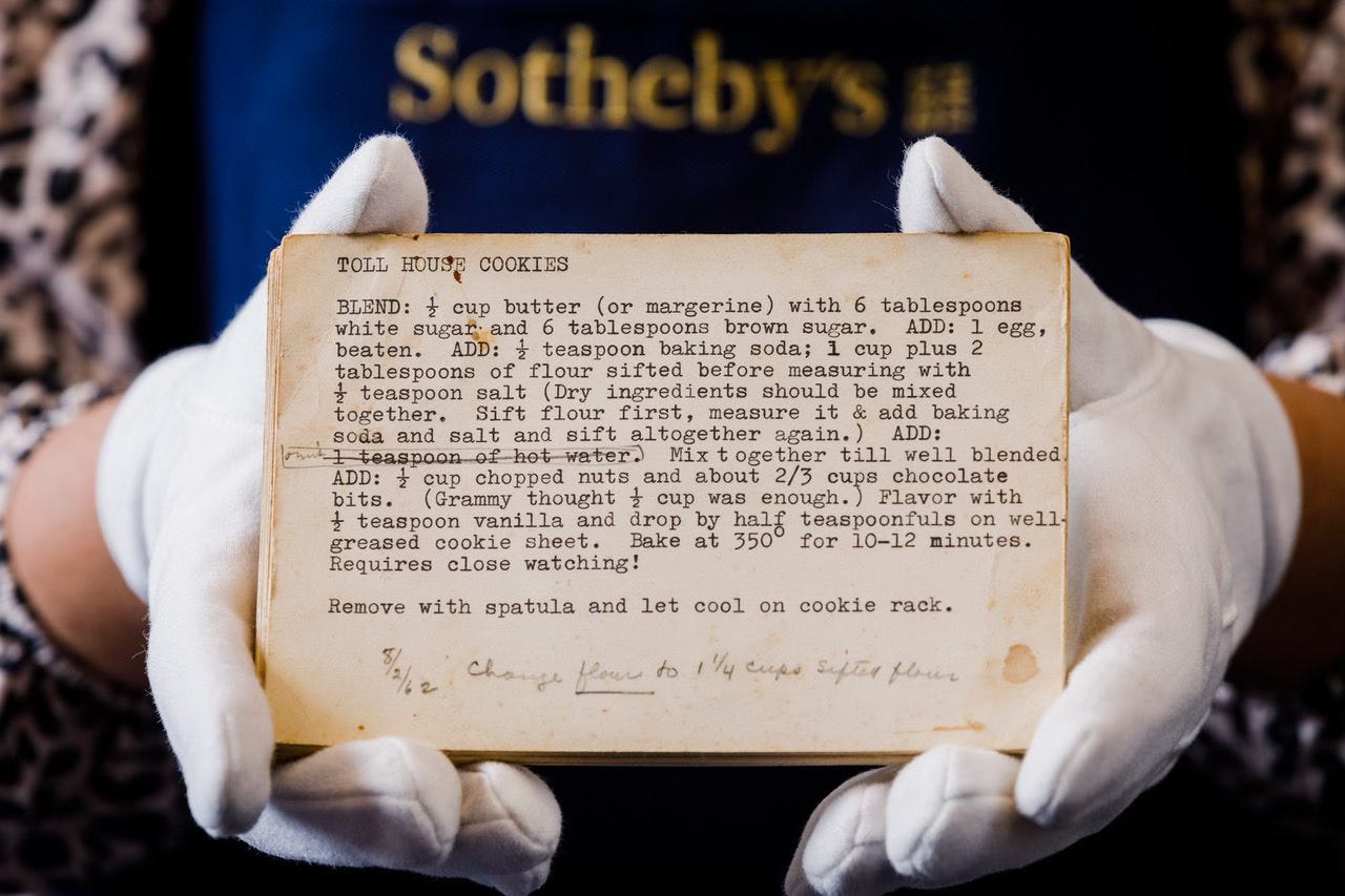 A typed recipe card with instructions for making Toll House Cookies, with handwritten corrections and additions in pencil. The card is held in gloved hands by a person wearing a Sotheby's apron.