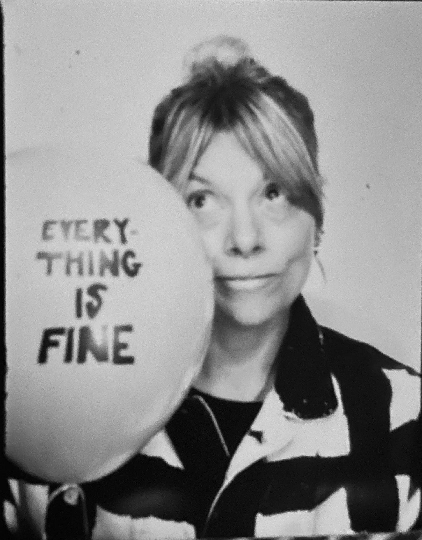 A black-and-white portrait of a woman with blonde hair tied up, wearing a patterned shirt and looking towards the camera with a slight smile. She holds a large balloon beside her face bearing the handwritten words "EVERYTHING IS FINE." A black-and-white portrait of a woman with blonde hair tied up, wearing a patterned shirt and looking towards the camera with a slight smile. She holds a large balloon beside her face bearing the handwritten words "EVERYTHING IS FINE."