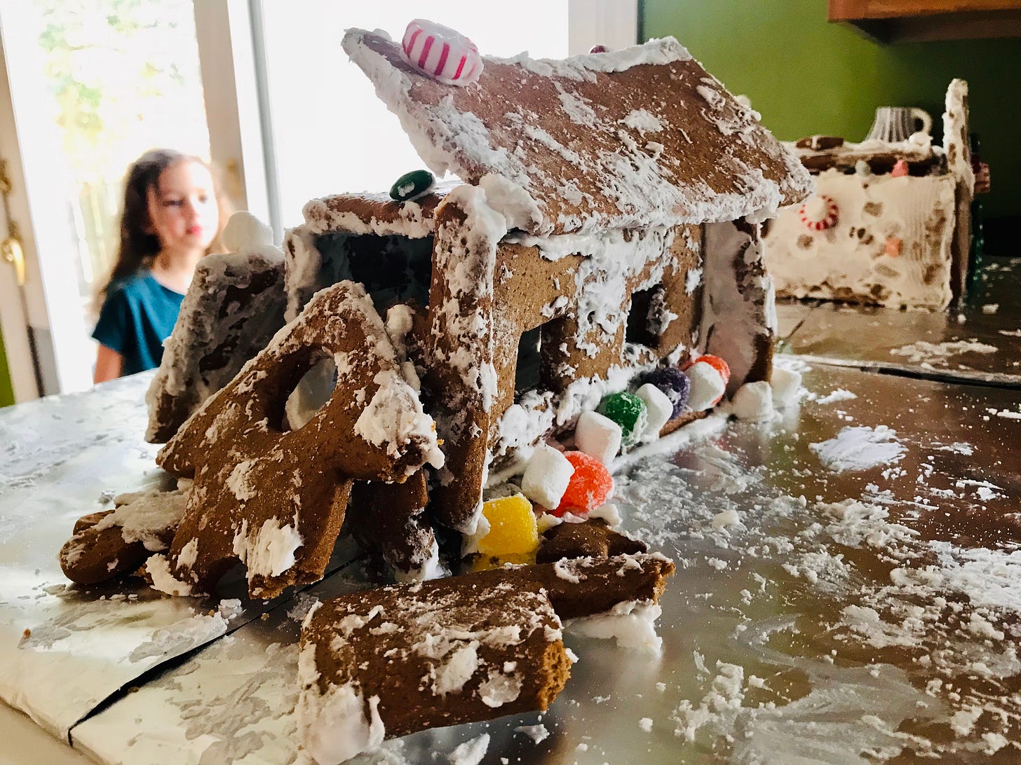 A gingerbread house collapsed on aluminum foil. Behind it, a blurry child and a backlit window.