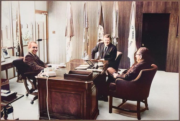 Henry Kissinger, Richard Nixon and John Wayne sit around a desk in an office in front of flags. Henry Kissinger, Richard Nixon and John Wayne sit around a desk in an office in front of flags.