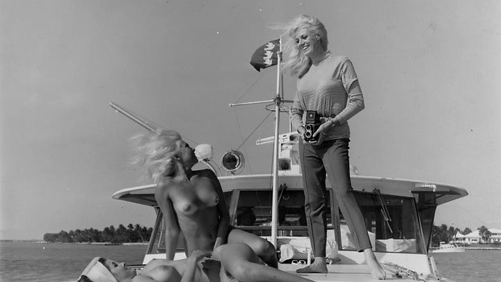 A four-image grid of vintage black-and-white photographs connected to Bunny Yeager: the top-left shows Yeager holding a camera while photographing a nude model lounging on the deck of a boat; the top-right shows a nude woman posing in a dramatic stance on an empty beach; the bottom-left shows a nude woman playfully pulling on a rope at the shoreline; and the bottom-right shows a woman in a bubble bath turning toward the camera with a smile.