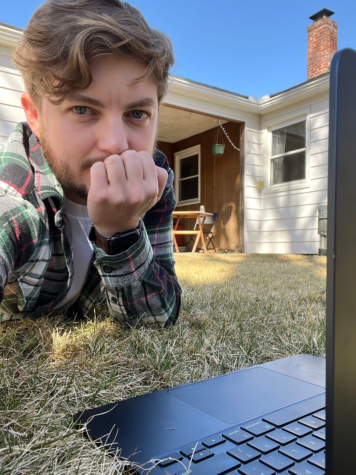 Jackson Bird lying on his stomach in the grass of his backyard, with his laptop open on the grass in front of him. He's looking straight into the camera as he takes a selfie.