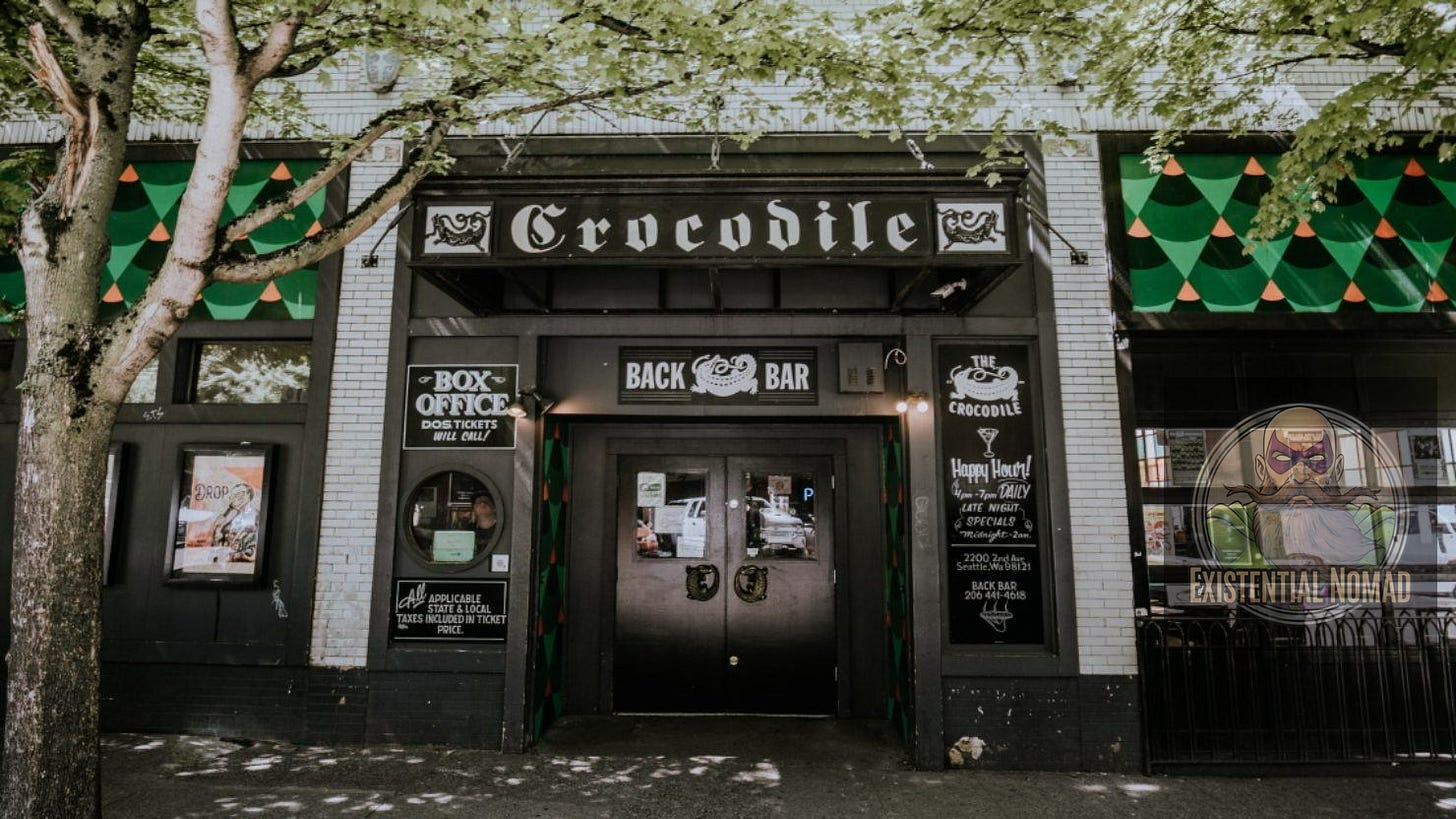 This is a photograph of the front entrance of a music venue called "The Crocodile." The building's facade is a mix of white and black tiles. A large, ornate sign over the main door reads "Crocodile." Below it, a sign for the "BACK BAR" and the main entrance doors are visible. The windows on either side are decorated with a green and orange geometric pattern and have a stylized, snake-like design painted on them. A tree with green leaves partially obstructs the left side of the image. The venue also has signs for "BOX OFFICE" and "Happy Hour!"