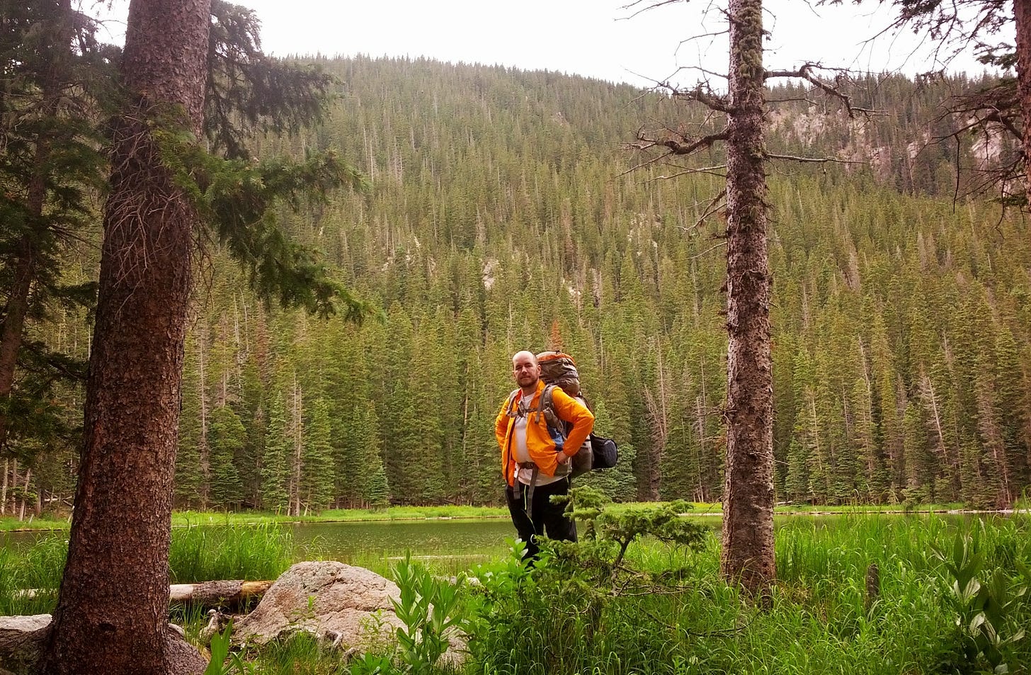 A man wearing an orange jacket with a fully loaded hiking packpage stands between trees near a lake. A small peak is in the near background.