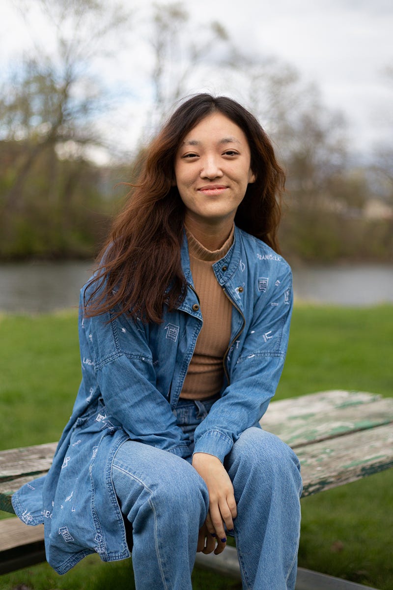 Portrait of Miri, a young, Asian-American woman with long brown hair. She is sitting on a picnic table at a park, smiling at the camera. She is wearing a denim overcoat with white-embroidered symbols and denim pants, with a brown, ribbed turtle neck underneath. Portrait of Miri, a young, Asian-American woman with long brown hair. She is sitting on a picnic table at a park, smiling at the camera. She is wearing a denim overcoat with white-embroidered symbols and denim pants, with a brown, ribbed turtle neck underneath.