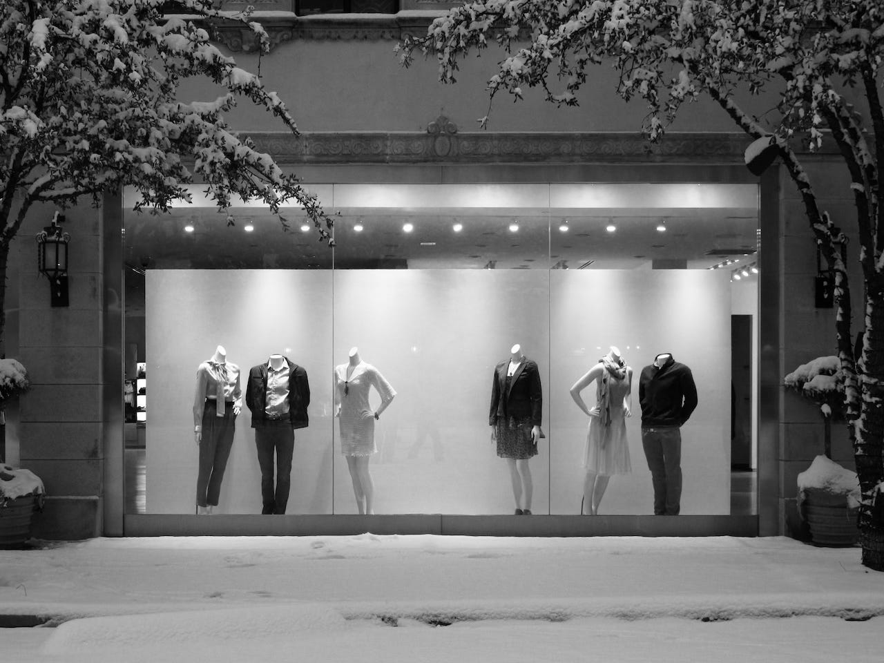 Black-and-white photo of a clothing store window display at night, with six headless mannequins dressed in winter and formal outfits. Snow covers the sidewalk and the branches of trees framing the storefront, while bright interior lights contrast with the quiet, snowy street outside. Black-and-white photo of a clothing store window display at night, with six headless mannequins dressed in winter and formal outfits. Snow covers the sidewalk and the branches of trees framing the storefront, while bright interior lights contrast with the quiet, snowy street outside.