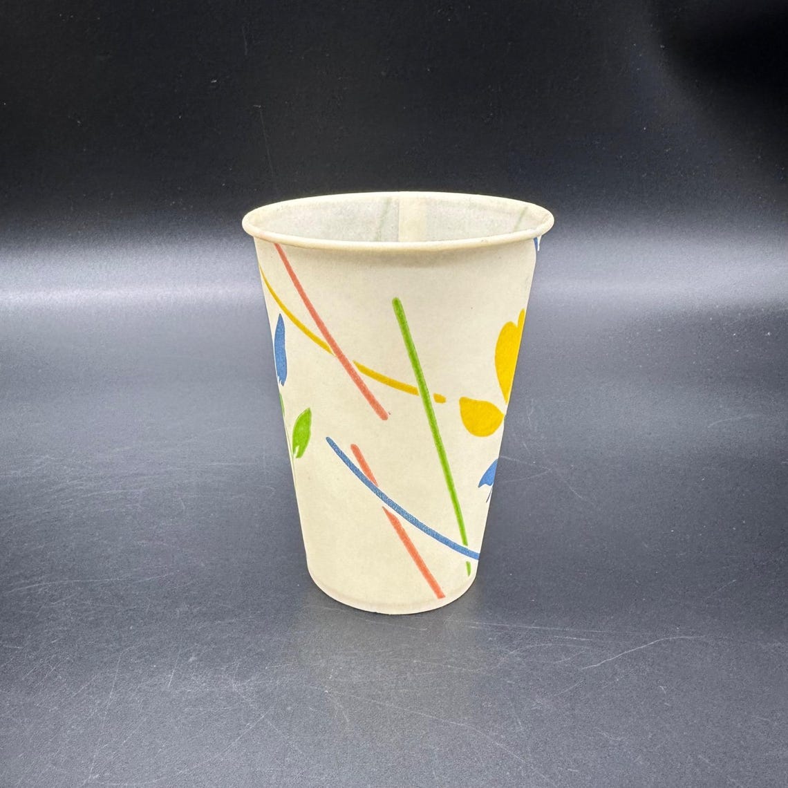 May include: A paper cup with a cream-colored background and colorful abstract floral and line designs in yellow, blue, red, and green. The cup is cylindrical with a slightly flared rim. May include: A paper cup with a cream-colored background and colorful abstract floral and line designs in yellow, blue, red, and green. The cup is cylindrical with a slightly flared rim.