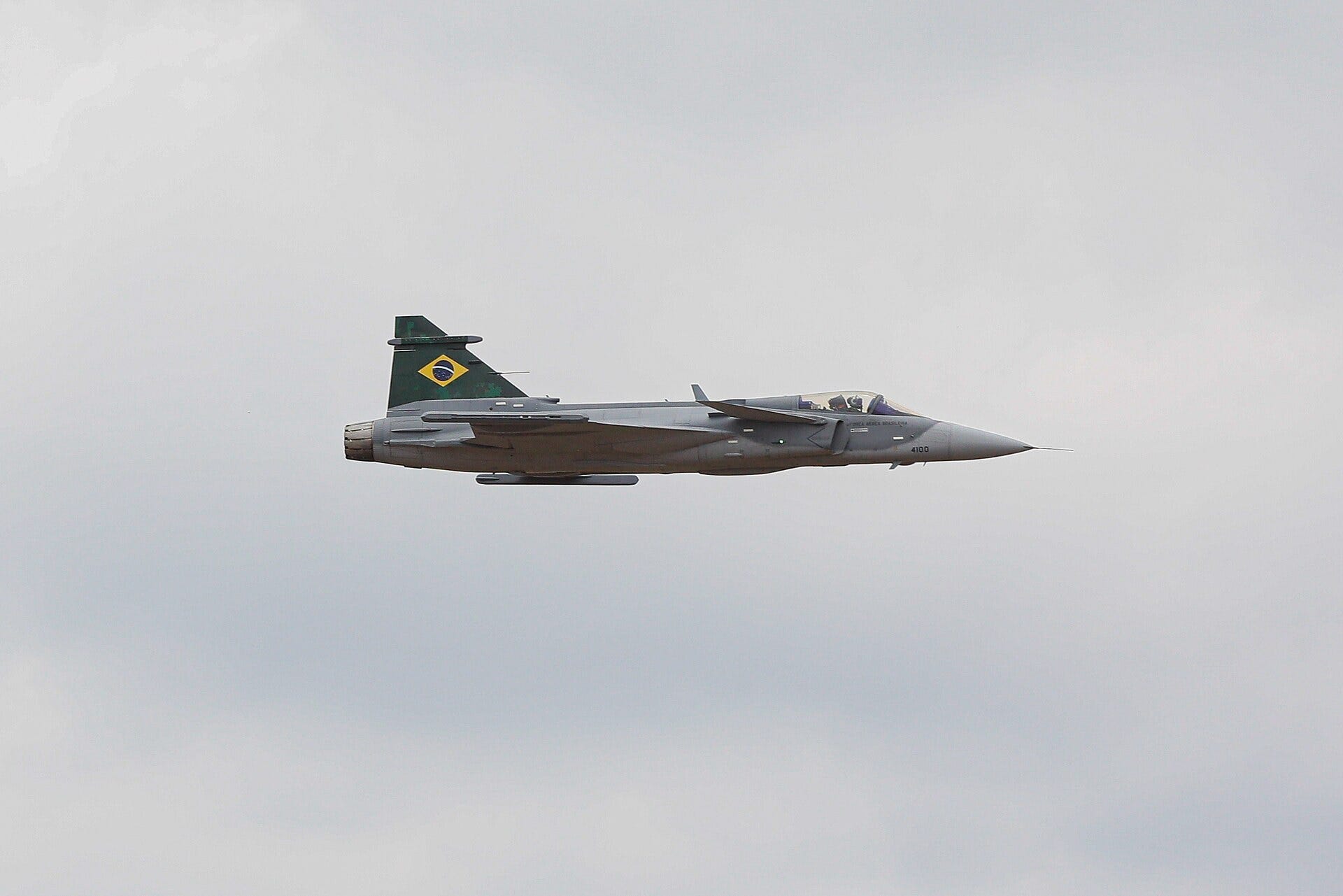 A grey F-39E Gripen with a Brazilian flag on its tail flying across a pale overcast sky.