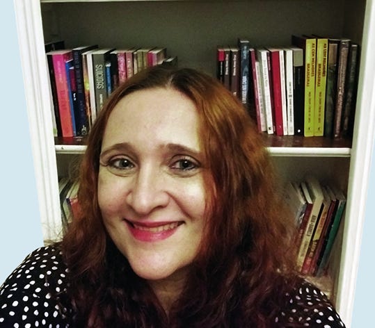Pessoa com cabelo comprido castanho-avermelhado, óculos, usando blusa preta com poás brancos, sorrindo para a câmera em primeiro plano, com estante de livros ao fundo contendo volumes coloridos organizados em prateleiras. Pessoa com cabelo comprido castanho-avermelhado, óculos, usando blusa preta com poás brancos, sorrindo para a câmera em primeiro plano, com estante de livros ao fundo contendo volumes coloridos organizados em prateleiras.