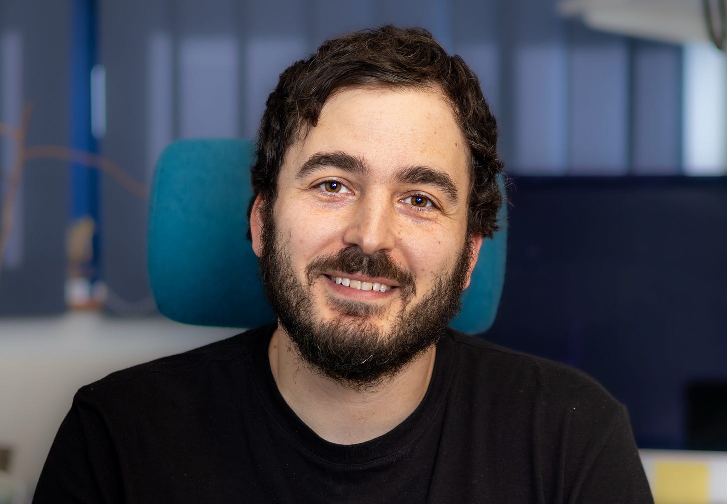 Image shows a male with brown hair and a beard wearing a black top and smiling at the camera