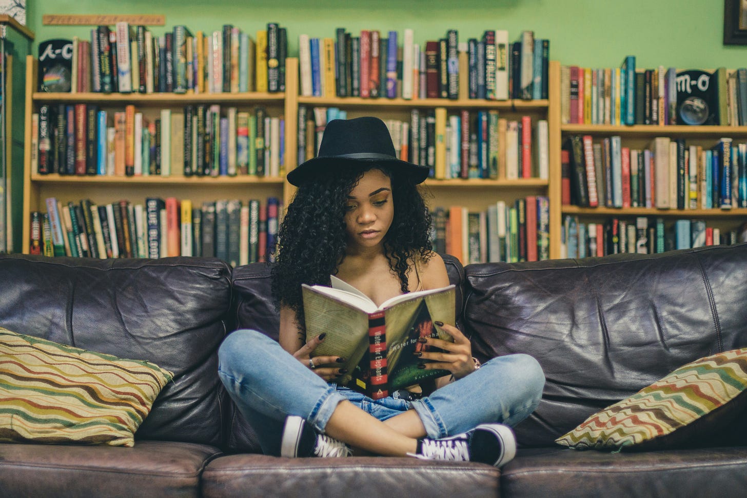 A black woman sitting cross-legged on a leather couch reading with bookshelves behind her A black woman sitting cross-legged on a leather couch reading with bookshelves behind her