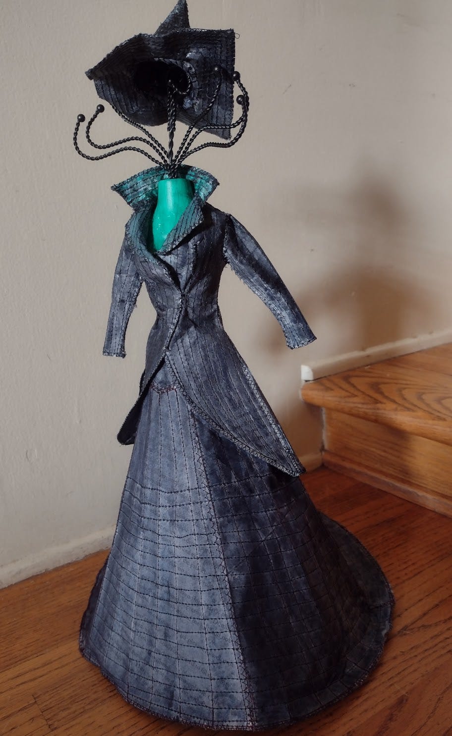 Green mini mannequin in a black paper tailcoat, skirt, and hat
