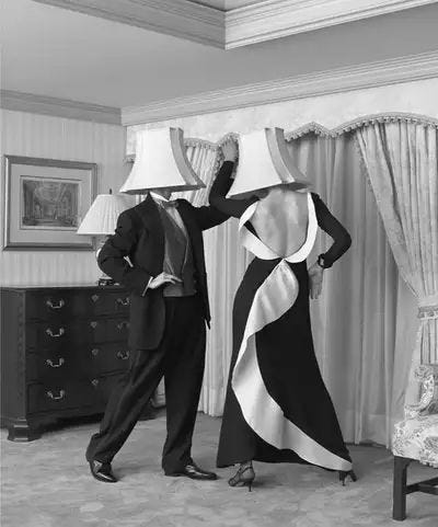 A black-and-white photo of two elegantly dressed people in a formal living room, each wearing a lampshade over their head. The person on the left is in a tuxedo, and the person on the right wears a floor-length evening gown with a dramatic white sash detail down the back. They appear to be mid-dance, striking a playful pose. A black-and-white photo of two elegantly dressed people in a formal living room, each wearing a lampshade over their head. The person on the left is in a tuxedo, and the person on the right wears a floor-length evening gown with a dramatic white sash detail down the back. They appear to be mid-dance, striking a playful pose.