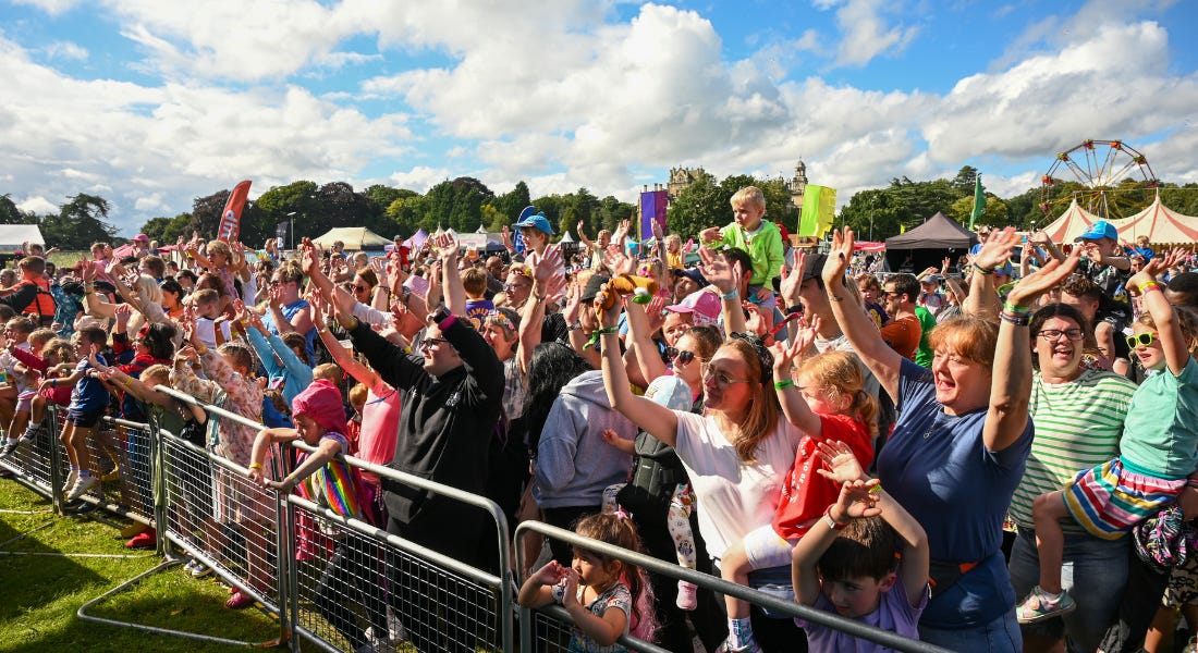 A crowd of people next to the railings of a stage at Gloworm Festival standing on grass, with a blue sky above them