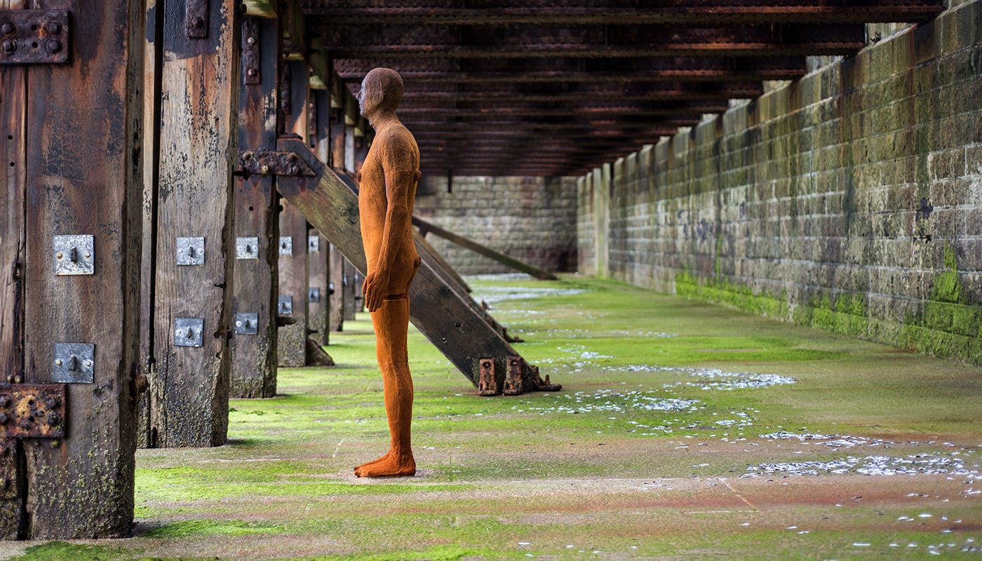 A statue of a man stands in profile as if looking out between some wooden columns. It is outside on the seashore and the tide is out