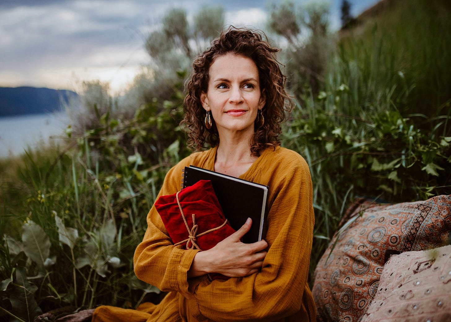 A woman stands calmly near a shoreline holding a book and a bound blanket. Photo by Amie Roussel on Unsplash
