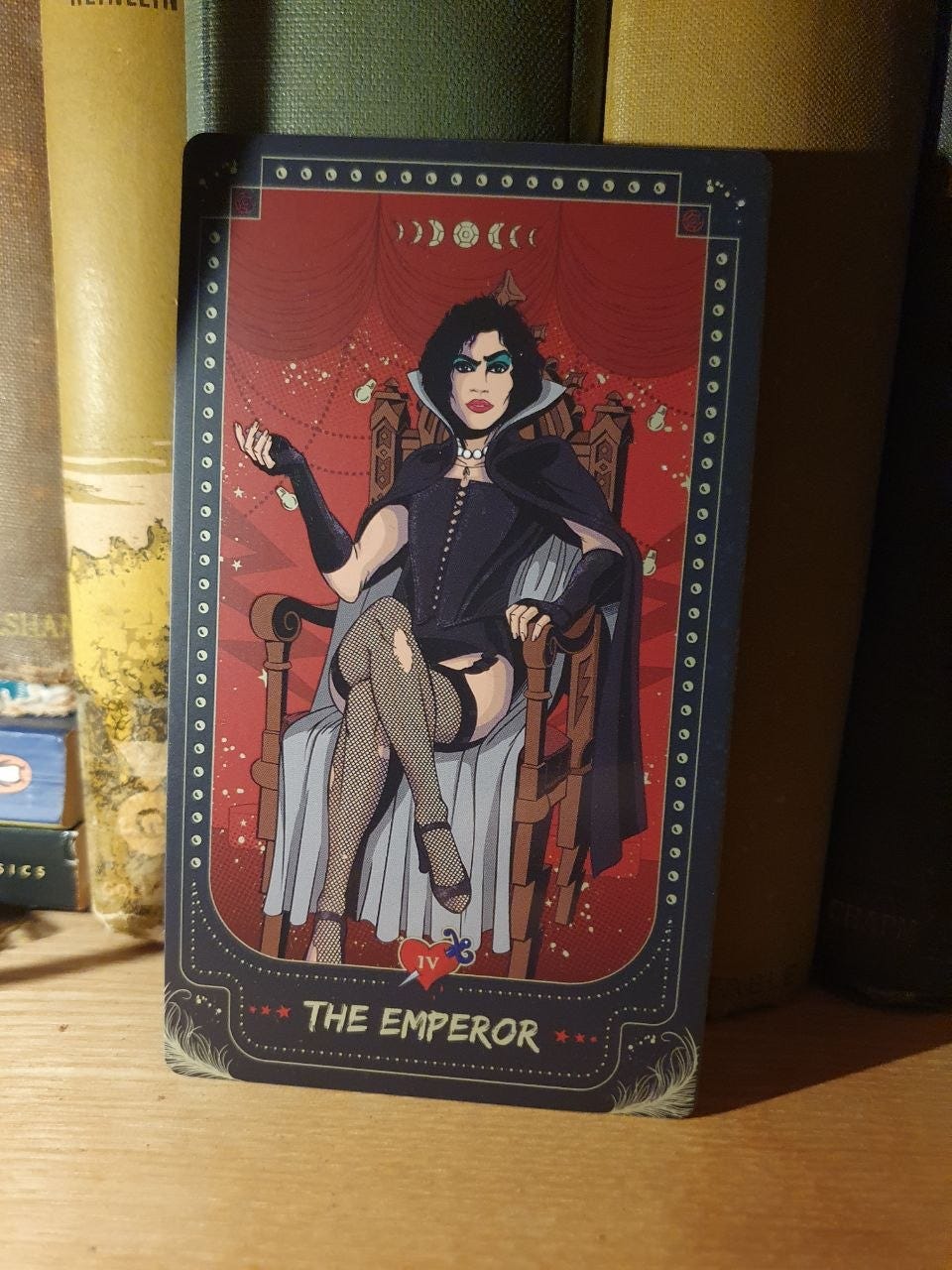 The Emperor tarot card, IV, featuring a dark-haired figure in a black cape, corset and fishnet stockings, seated on an ornate wooden throne against a red theatrical backdrop. The Emperor tarot card, IV, featuring a dark-haired figure in a black cape, corset and fishnet stockings, seated on an ornate wooden throne against a red theatrical backdrop.
