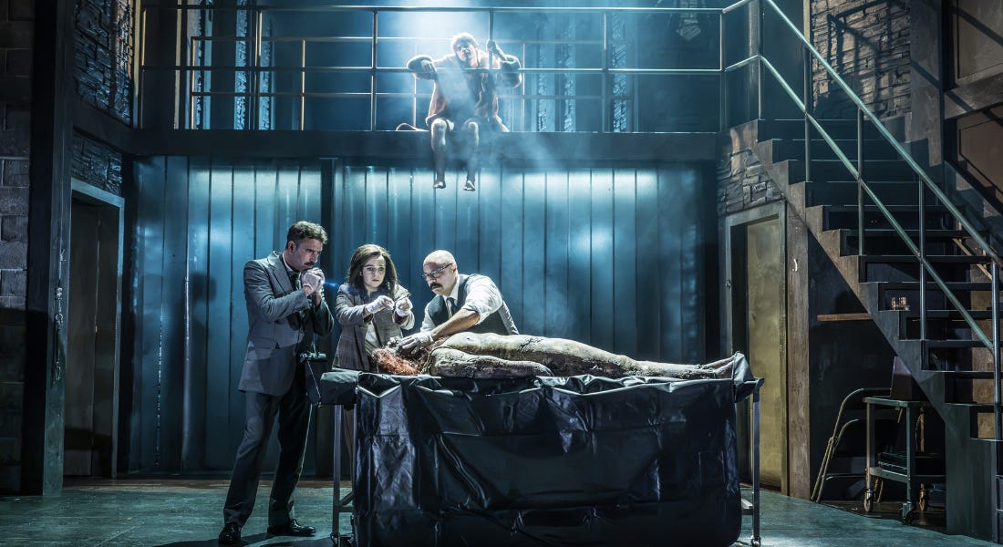 A group of people stood around a table with a corpse, with a man sat on the balcony above