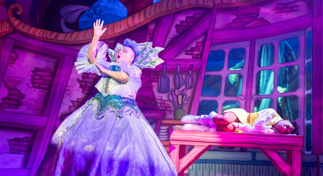 A woman, in a sparkling blue dress with a hooped skirt, stands in a storybook-style kitchen on a stage. She is signing the word ‘beanstalk’ in BSL and singing. On a table behind her, a man is curled up asleep