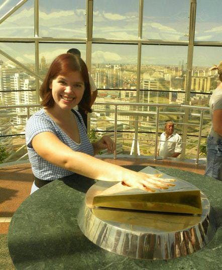 Katharine Duckett, pictured here in her early 20s, is a white woman with red hair, smiling and wearing a blue short-sleeved checkered dress. She has her hand on a golden triangle in the windowed viewing platform atop Bayterek Tower in Nur-Sultan, the capital of Kazakhstan.