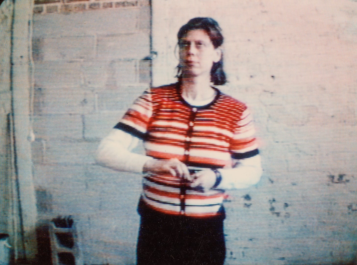 Lo-res video still of the artist, Mary Addison Hackett standing in an empty room facing the camera. She prepares to throw a dart. She’s wearing a pink, white, and black striped shirt and black pants. She is rolling her eyes. Lo-res video still of the artist, Mary Addison Hackett standing in an empty room facing the camera. She prepares to throw a dart. She’s wearing a pink, white, and black striped shirt and black pants. She is rolling her eyes.