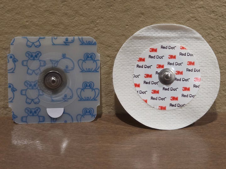 Photos of 3 different types of cardiac electrodes, the sticky pads used on Holter monitors and other devices used to check for arrhythmias. There is one set of 3 foam electrodes with a lot of gel in the center; one “Huggable” children’s electrode that is plastic; and one 3M electrode that is cloth with a small amount of gel in the center.