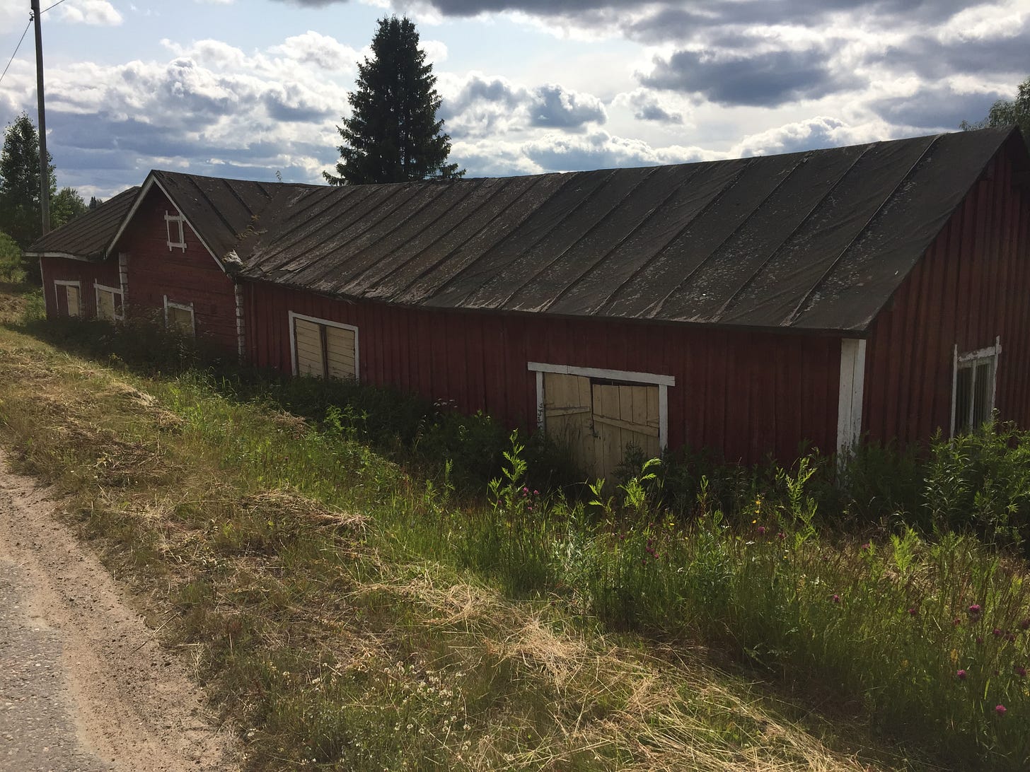 A long, low, old, wooden building, painted dark red, against a bright sky with clouds.