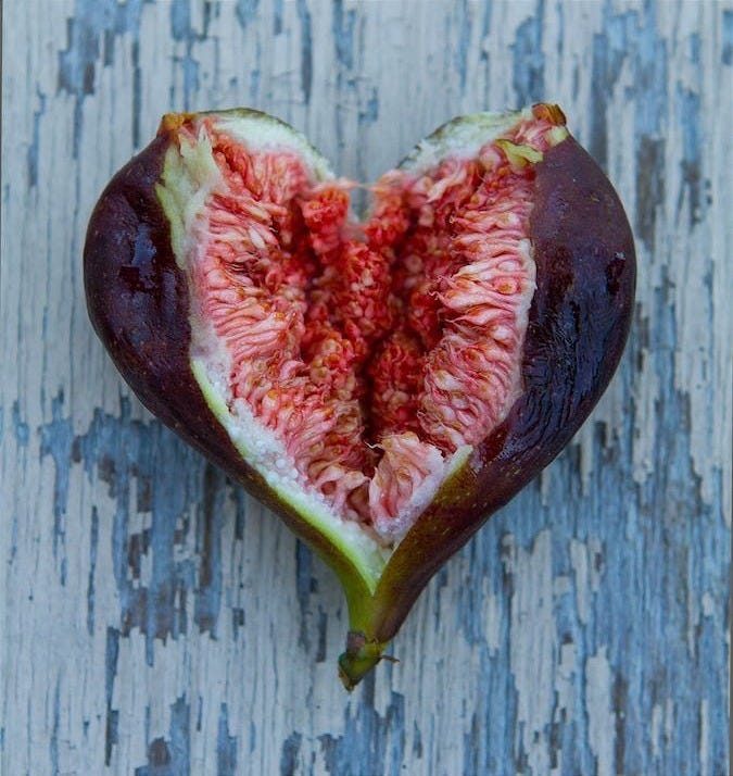 A ripe fig, split open at the centre, sits on weathered pale wood. Its rich red interior spills outward in fibrous folds, suggestive and raw, shaped into something that resembles both a heart and a vulva.